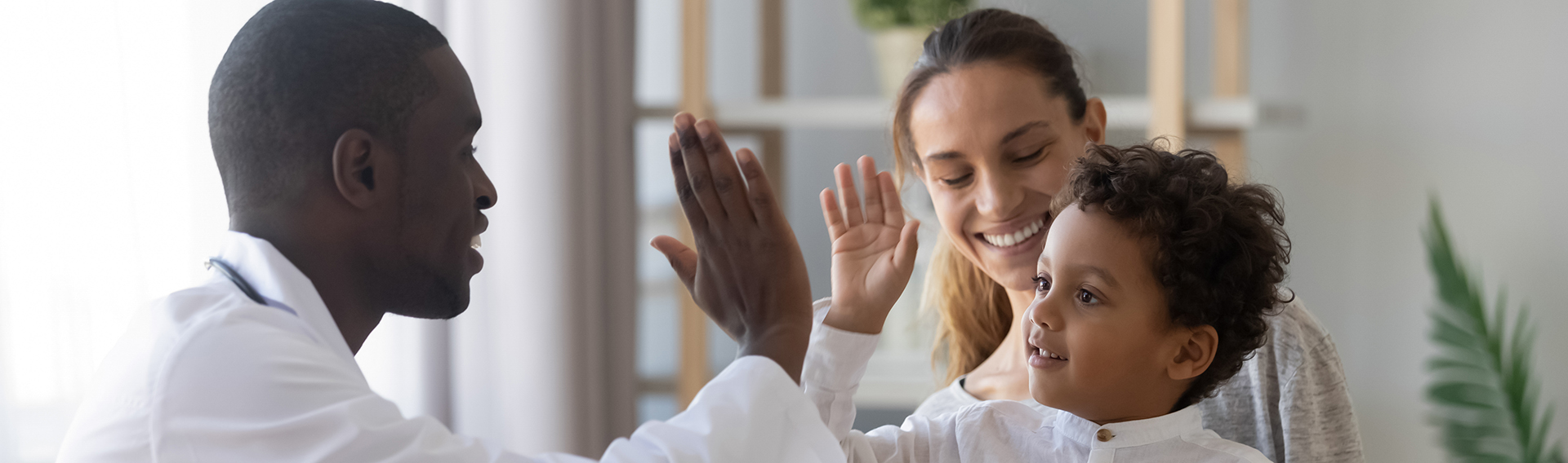 Little boy sit on mom laps give high five to African doc in white coat during visit in clinic.